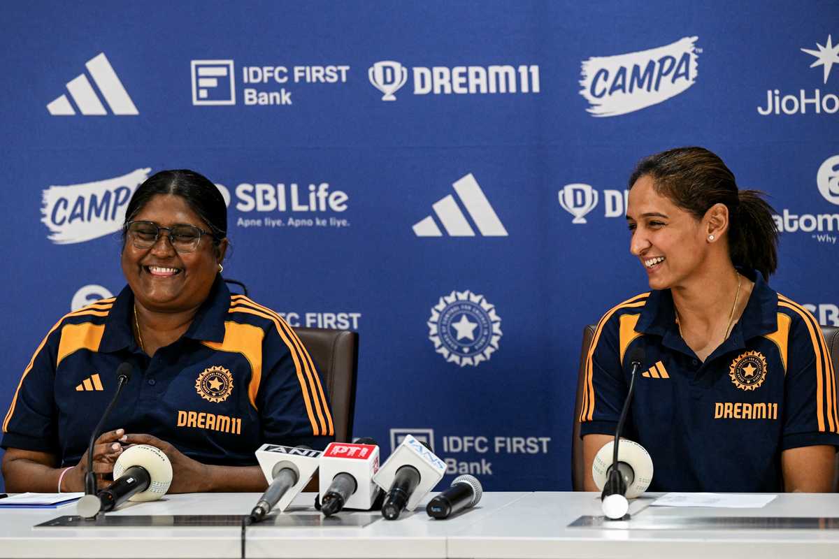 Two women smiling at a sports press conference, wearing matching team jerseys.