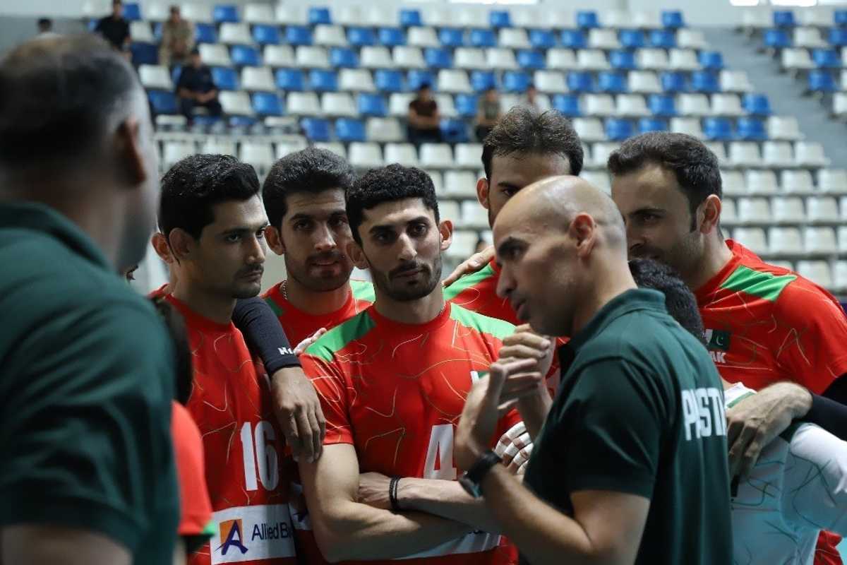 Volleyball team huddled, wearing red jerseys, in a sports arena.