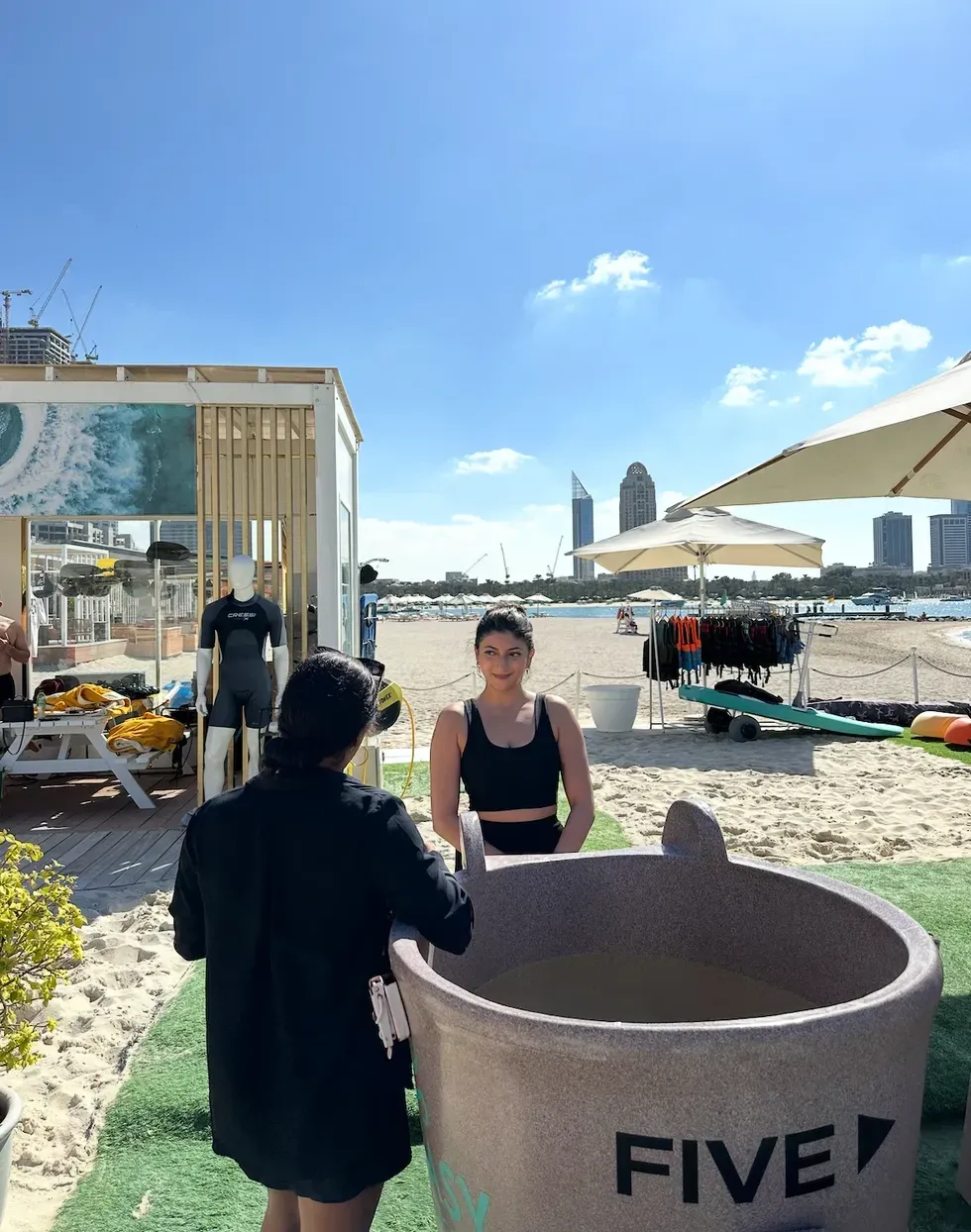 Woman in black top entering a large tub on a sunny beach with city skyline in background.